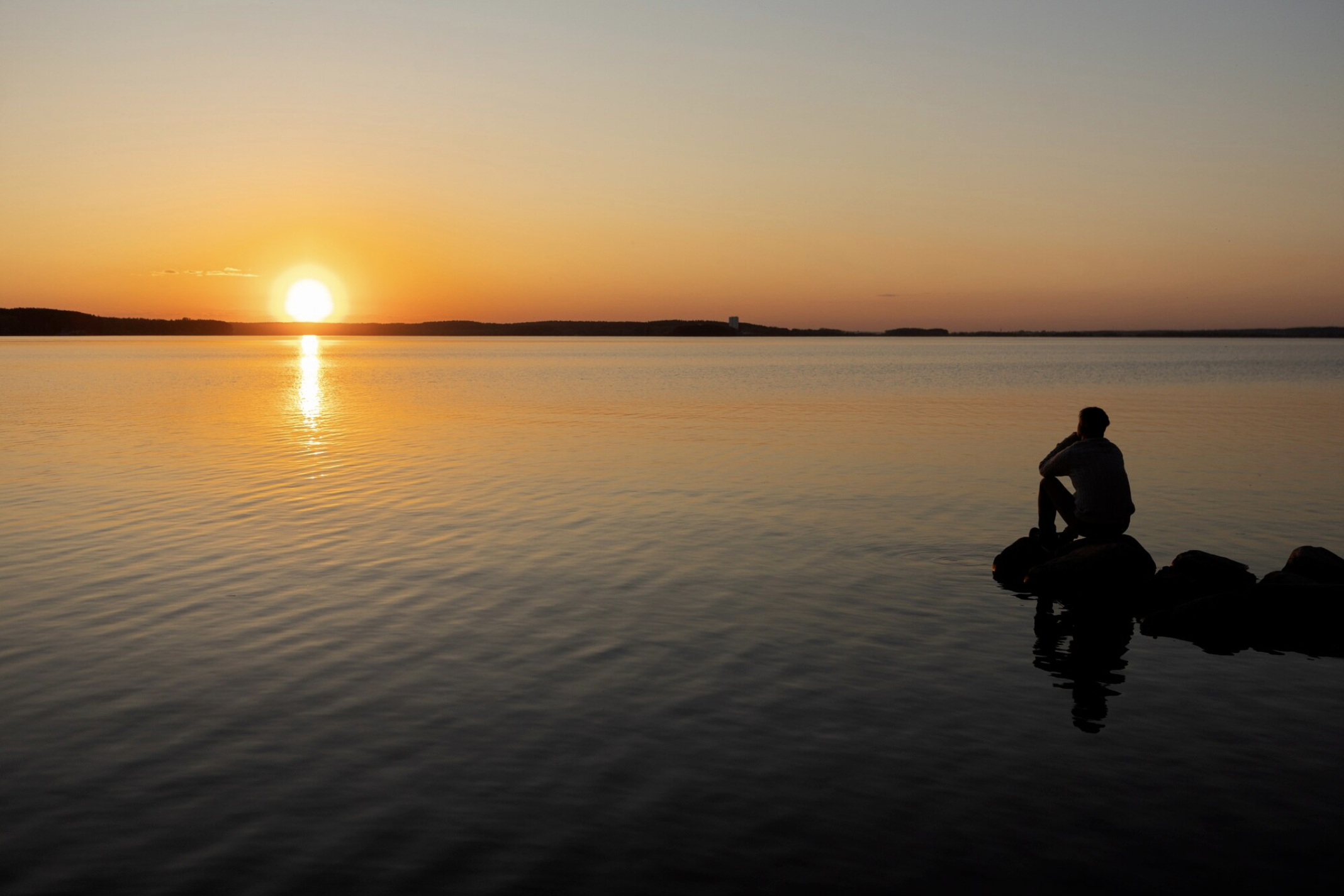 Ein Mann der am See trauert. Er sitzt auf einem Felsen und schaut in den Sonnenuntergang.